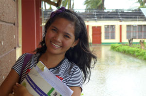 Mirinel är 17 år och lever i Filippinerna. Hon håller en bok i famnen och står framför en översvämmad gata.