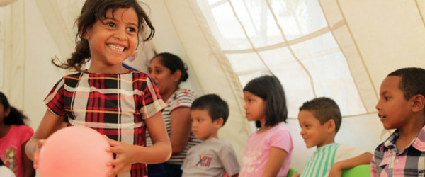 En flicka i förgrunden ler och håller i en ballong. I bakgrunden står barn och lärare. 