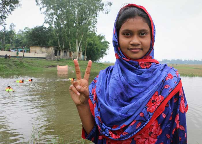 10-åriga Irin i Bangladesh lär yngre barn simma. På bilden står hon framför ett vattendrag, tittar in i kameran och gör fredstecknet med fingarna.