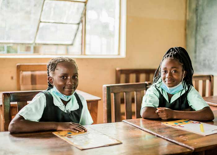 Två flickor sitter i ett klassrum i Zambia med skolböcker framför sig på bänkarna och munskydd hängande runt halsen. 