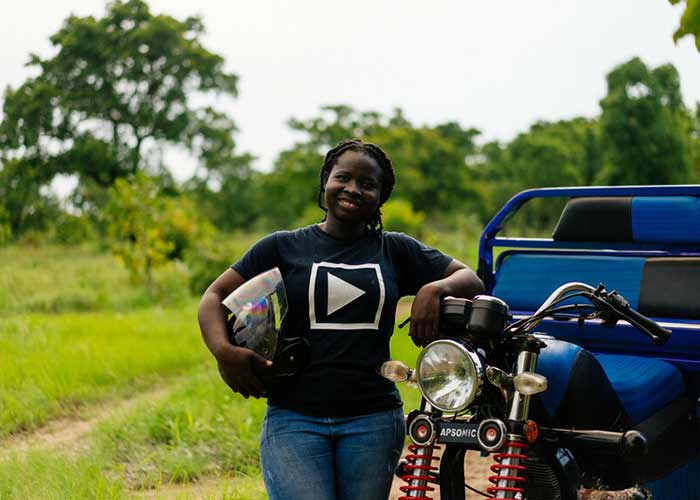 24-åriga Olivia tittar leende in i kameran där hon står utomhus med armen lutad mot sin trehjuling.