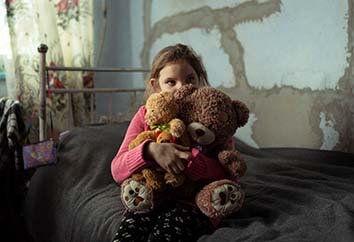 Flicka som flytt griget i Ukraina sitter på en säng och kramar sina nallebjörnar.