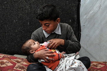 Pojke matar ett yngre barn i ett tillfälligt läger i Södra Gaza.